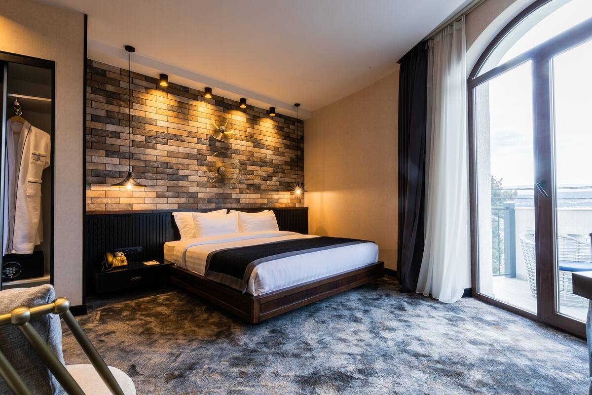 A modern hotel room with a brick-patterned accent wall, a large bed, a wardrobe, and a view of the outdoors.