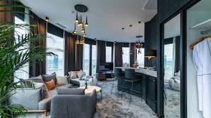 Un salon moderne avec un canapé gris, une table basse et un téléviseur. Une grande fenêtre, ornée de rideaux, laisse entrer la lumière et offre une vue sur la ville. On y aperçoit également une plante.