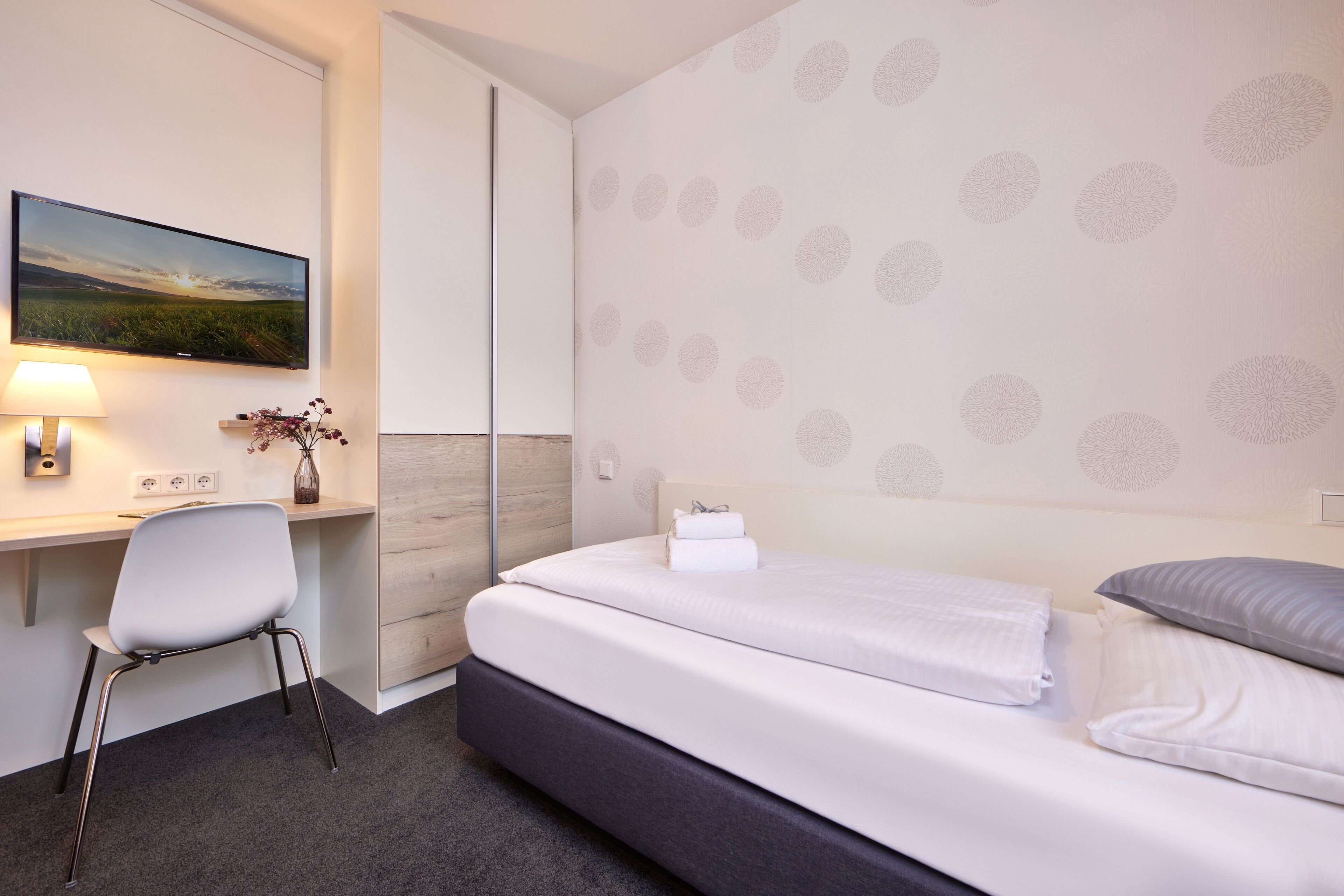 A hotel room with a bed, a desk, a chair, a TV, and a patterned wall.