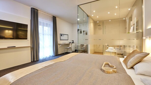 A modern hotel room with a large bed, a desk, a TV, and a glass-enclosed bathroom. - Hotel Alter Wirth (Geretsried)