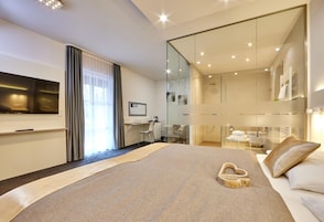A modern hotel room with a large bed, a desk, a TV, and a glass-enclosed bathroom.