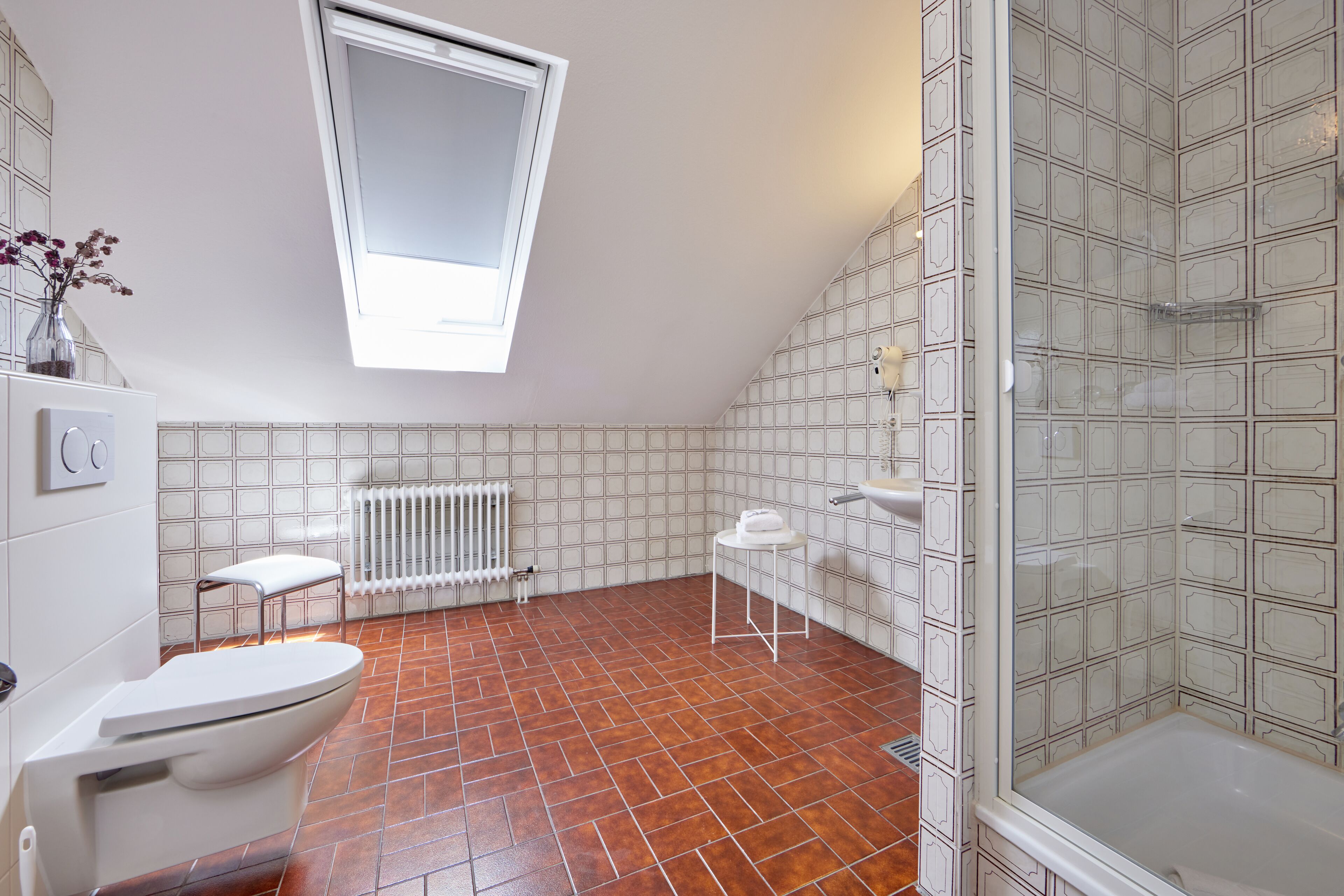 Um banheiro moderno com teto inclinado, claraboia, vaso sanitário branco, chuveiro com box de vidro e piso revestido de azulejos.
