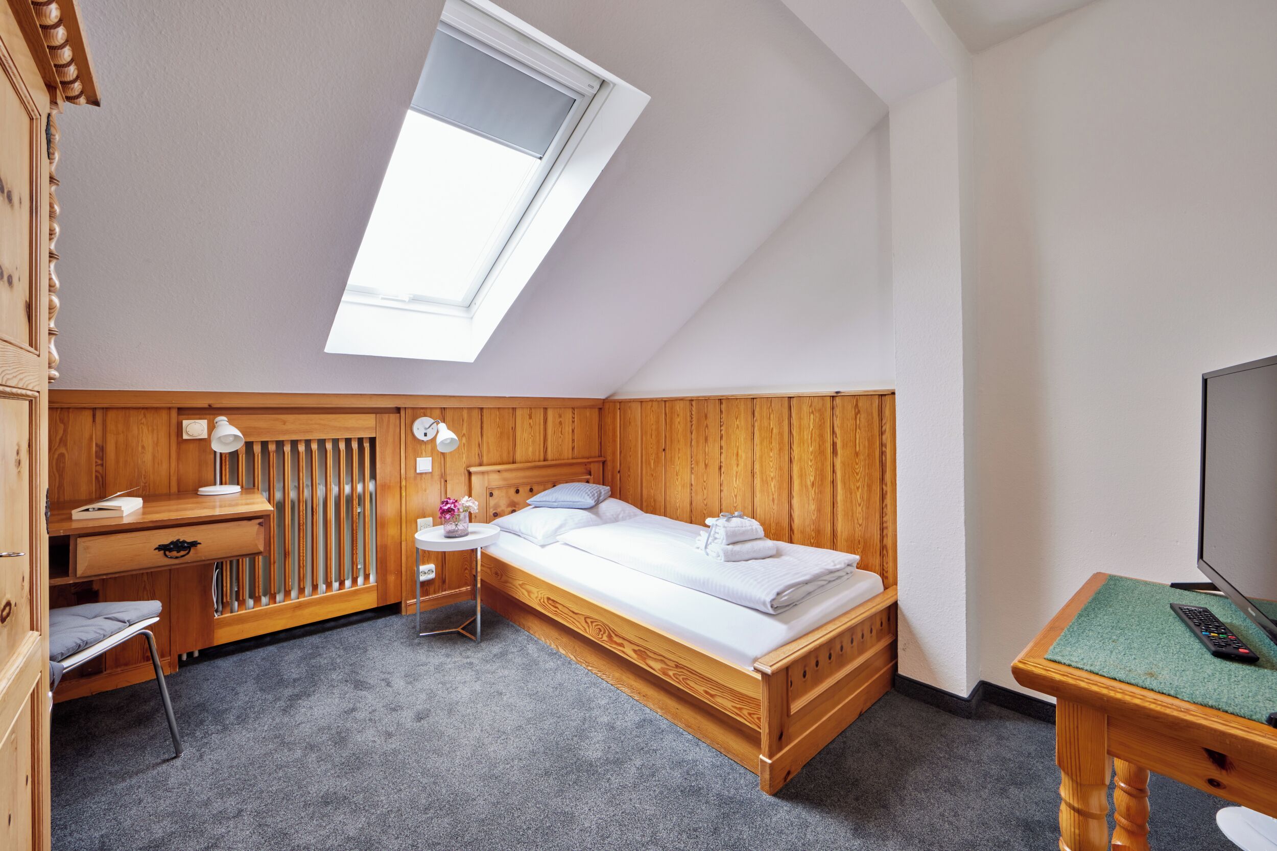 Une pièce avec un plafond en pente, un lit, un bureau, une chaise, une petite table et un téléviseur.