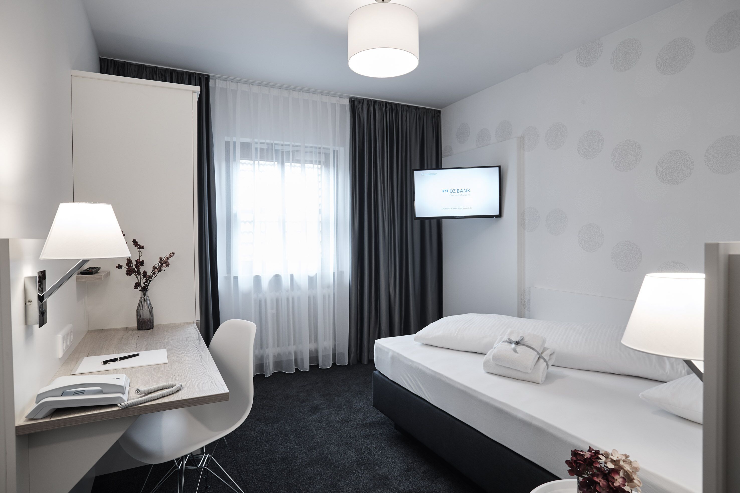Quarto de hotel com escrivaninha, cadeira, cama e televisão exibindo 'DZ BANK'.