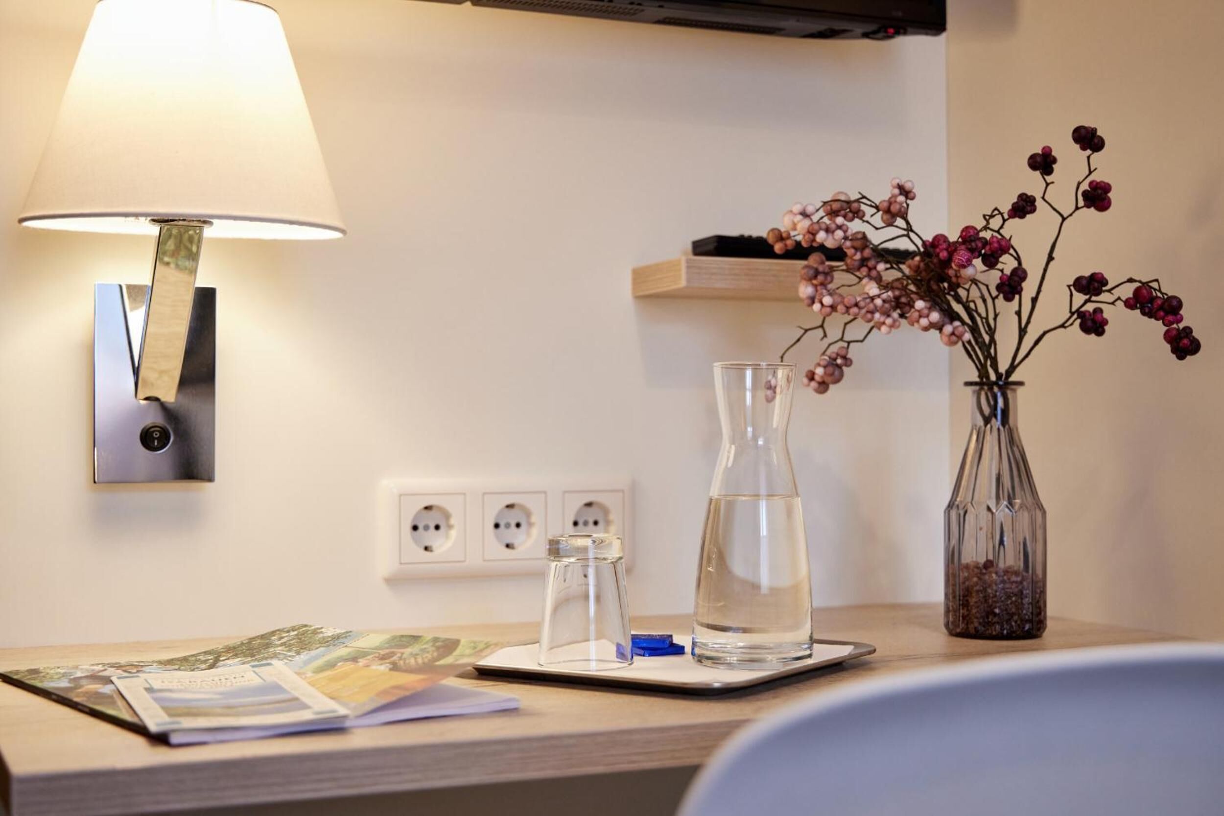 A neatly arranged desk with a lamp, a vase with flowers, a water bottle, and a glass jar.