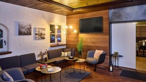A modern living room with a wooden ceiling, a large flat-screen TV, and a cozy seating area with a sofa and armchairs. - Hotel Alter Wirth (Geretsried)