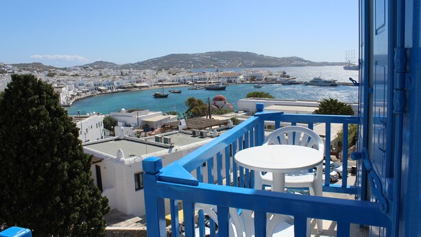 A balcony with a white table and chairs, overlooking a coastal town with boats and buildings. - Stelios View Mykonos Town (Mykonos)