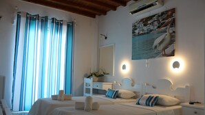 A hotel room with two beds, a white dresser, a large window with curtains, a painting of a pelican, and wall-mounted lights. - Stelios View Mykonos Town (Mykonos)