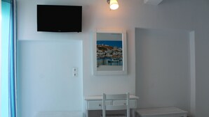 A white room with a desk, two chairs, a television, and a framed picture of a coastal town. - Stelios View Mykonos Town (Mykonos)