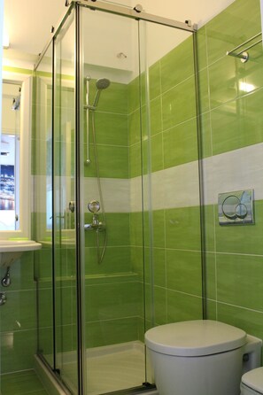 A bathroom with a glass-enclosed shower, green tiles, a white toilet, and a sink. - Stelios View Mykonos Town (Mykonos)