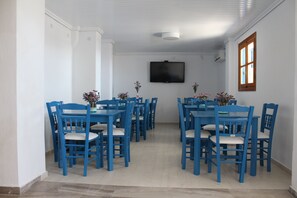 A dining area with blue tables and chairs, a flat-screen TV mounted on the wall, and a window with wooden shutters. - Stelios View Mykonos Town (Mykonos)