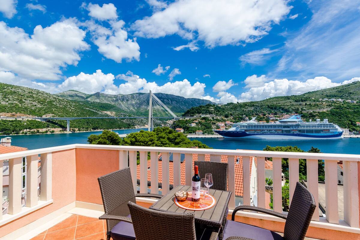 A balcony with wicker chairs and a table, overlooking a coastal area with a bridge and a cruise ship.