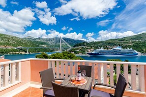 A balcony with wicker chairs and a table, overlooking a coastal area with a bridge and a cruise ship. - Guest House Avdic (Dubrovnik)