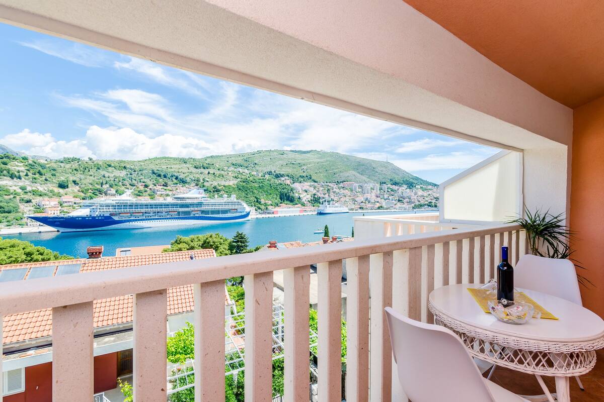 A balcony with a view of a cruise ship on a river, a table with a bottle, and a chair.