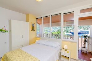 A bedroom with a bed, bedside table, lamp, and a window with a view of greenery and buildings. - Guest House Avdic (Dubrovnik)