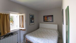 Un dormitorio con cama, ventana con cortinas, radiador y dos cuadros en la pared.