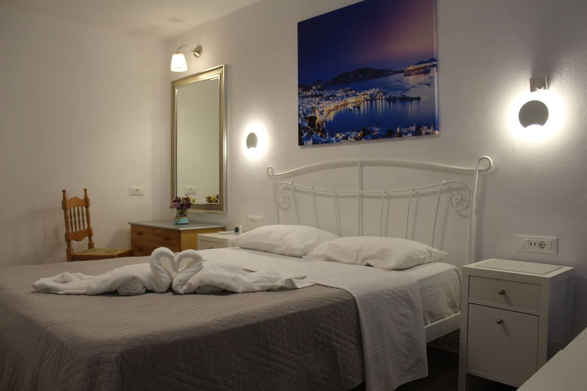 Una cama bien hecha con edredón blanco y almohadas, una silla de madera, una mesita de noche con lámpara, un mueble auxiliar con cajón y un espejo montado en la pared.