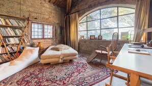 A rustic room with a large wooden beam ceiling, a bookshelf, a cushioned bench, a patterned rug, and a large arched window. - Bomarzo Ancient Villa (Bomarzo)