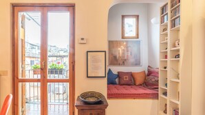 Una habitación acogedora con puerta de madera, una mesita con un cuenco, una estantería, un rincón con cojines junto a la ventana y un cuadro enmarcado en la pared.
