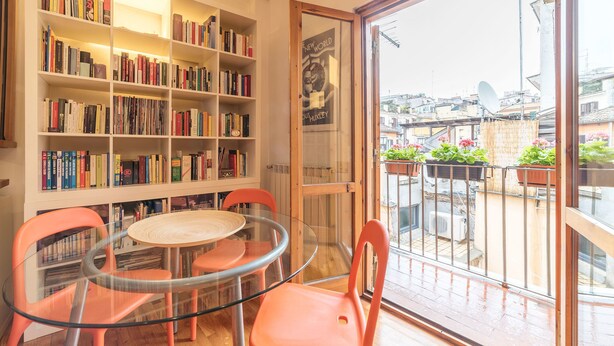 Una habitación con una mesa y sillas de vidrio, una estantería llena de libros y vistas a la ciudad a través de un ventanal grande.