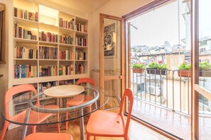 A room with a glass table and chairs, a bookshelf filled with books, and a view of the city through a large window. - Boschetto Peaceful Apartment (Rome)