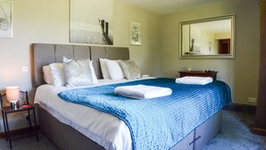 A neatly made bed with a blue blanket, a bedside table with a lamp, and a large mirror on the wall. - Idylic country cottage with hot tub on private estate (Challock)