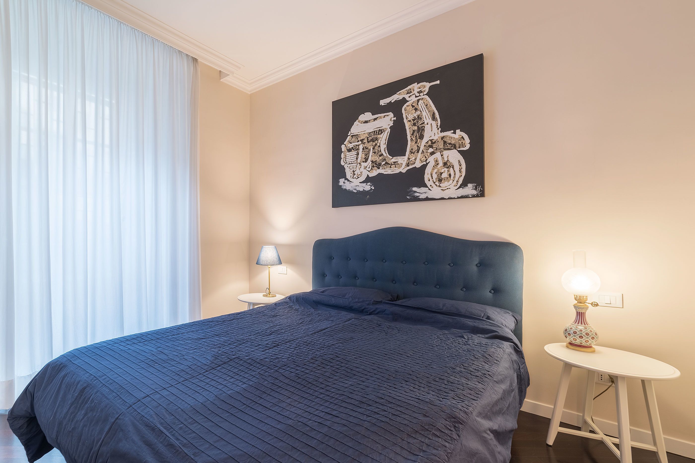 A bedroom with a large bed, a headboard, a wall-mounted artwork of a scooter, and a bedside table with a lamp.