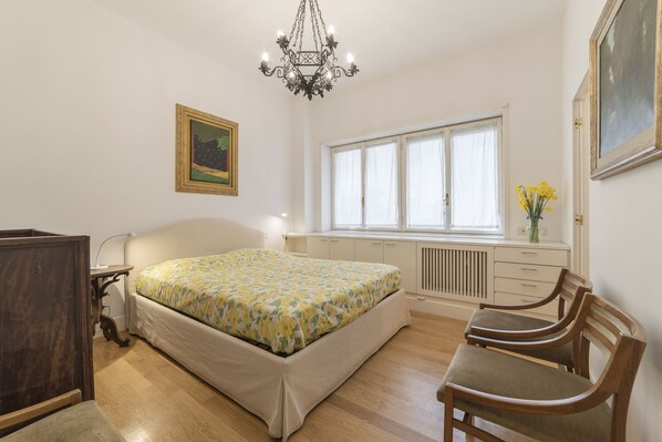 A bedroom with a bed, a wooden chair, a desk, a window, and a radiator.