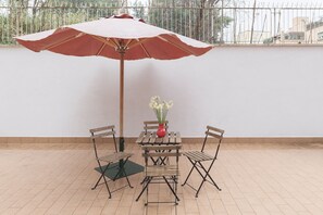 A patio with a table and chairs under a red umbrella, a vase with flowers, and a view of buildings in the background.