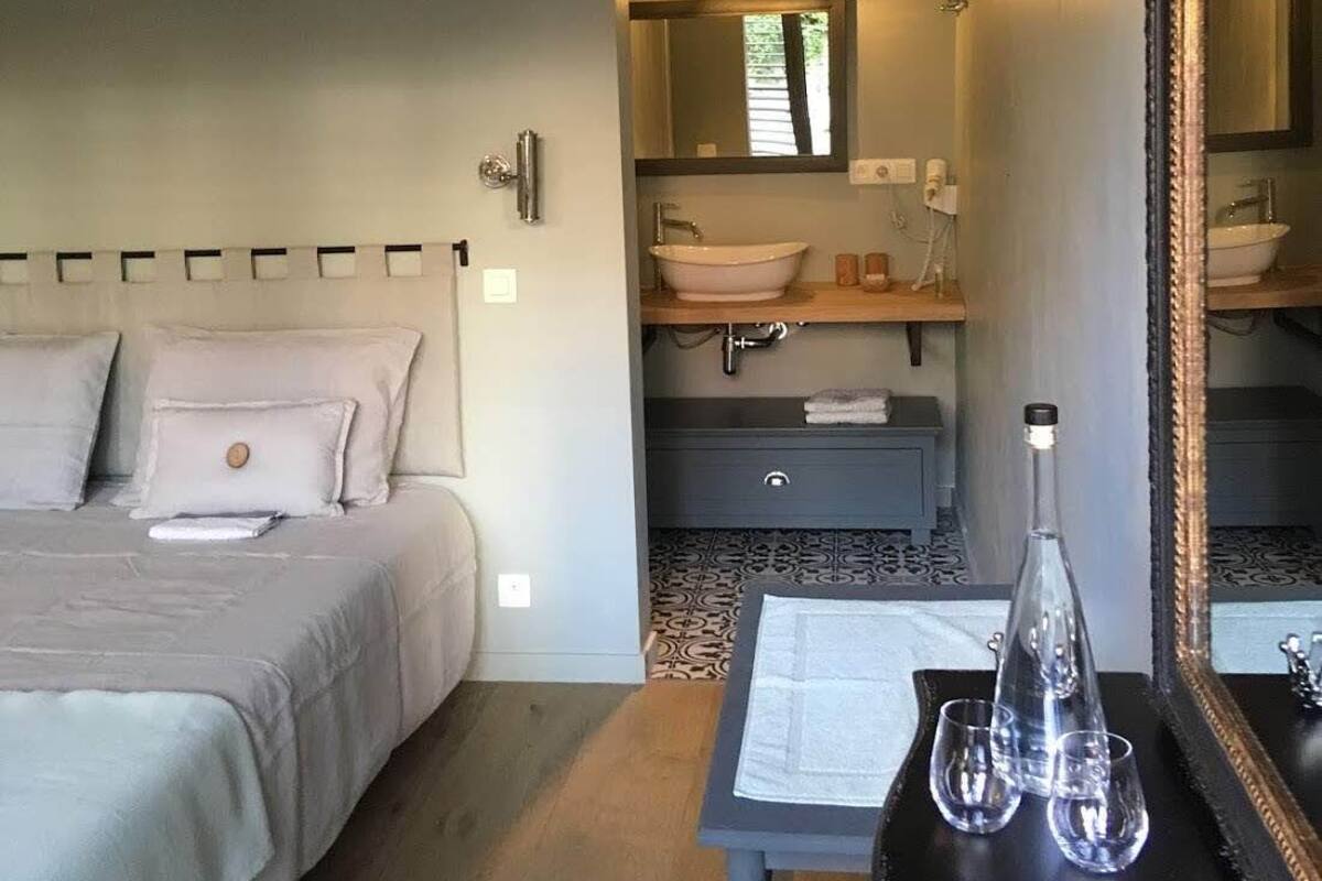 A bedroom with a bed, a bedside table with a bottle and glasses, and a bathroom with a sink and mirror.
