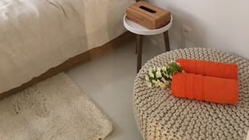 A bedroom with a bed, a small round stool, a white bedside table, and a white wall with a light switch.