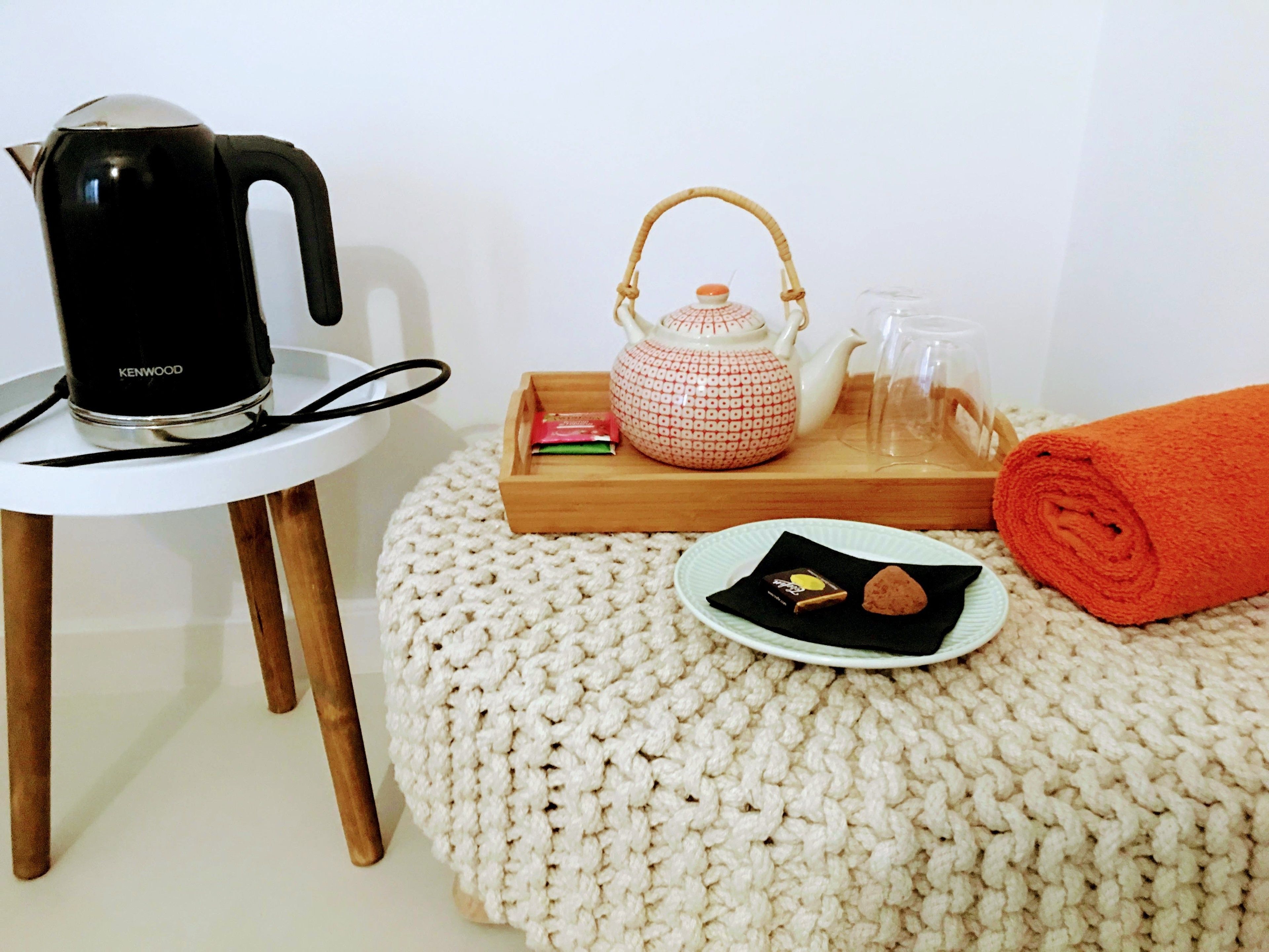 A cozy room with a knitted pouf, a white table with a black kettle, a wooden tray with a teapot, a glass pitcher, and a rolled orange towel.