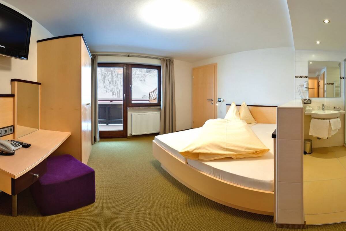 A hotel room with a bed, a desk with a telephone, a bathroom with a shower, and a TV mounted on the wall.