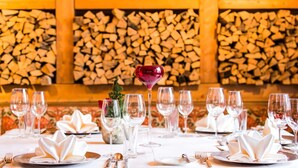 A dining table set for a meal with multiple wine glasses and plates, set against a backdrop of stacked firewood. - Hotel & Art Kristiana (Saalbach-Hinterglemm)