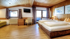 A hotel room with a wooden floor, a bed with pillows, a desk with a chair, a TV on the wall, and windows with curtains. - Hotel & Art Kristiana (Saalbach-Hinterglemm)