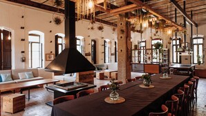Een ruime eethoek met een lange tafel, meerdere stoelen, hangplanten en een grote, zwarte schoorsteen.