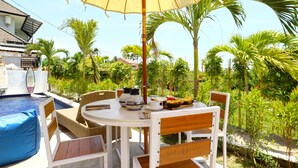 A round table with chairs and a yellow umbrella, surrounded by lush greenery and a swimming pool. - Margarita Surf Hostel Canggu (Pererenan)