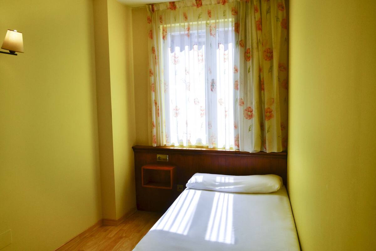 A room with a bed, a nightstand, a lamp, and a window with floral curtains.