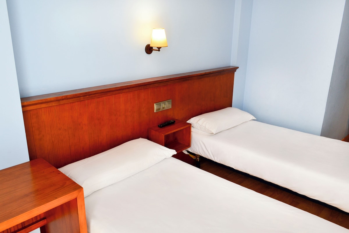A hotel room with two single beds, a wooden nightstand with a remote control, and a wall-mounted lamp.