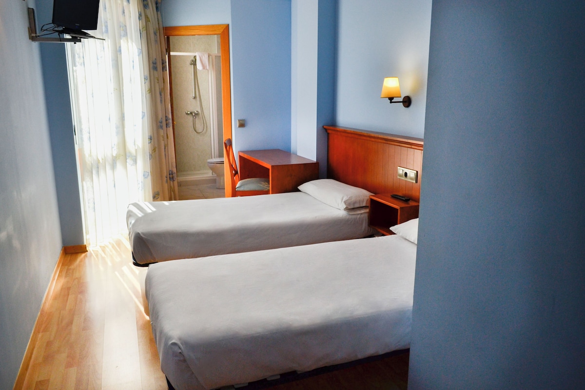 A hotel room with two single beds, a wooden nightstand, a small table, a TV mounted on the wall, and a shower area visible through an open door.