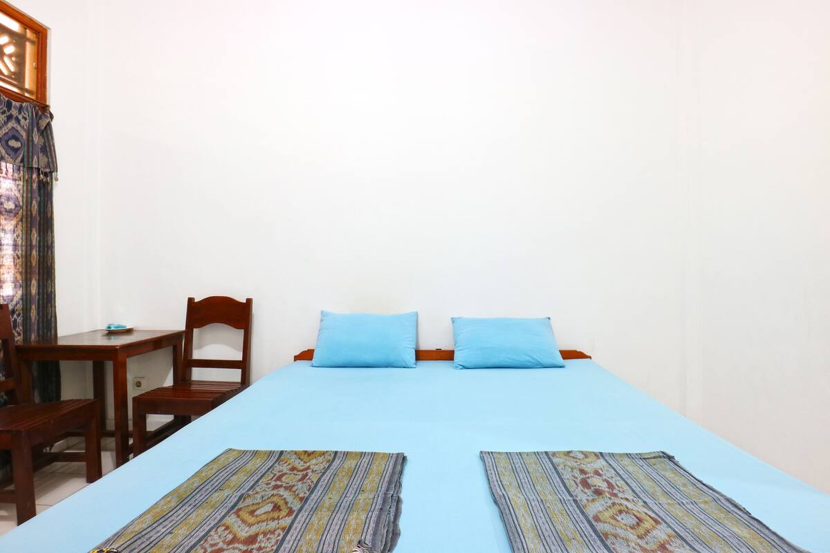 A room with a bed, two blue pillows, a wooden chair, a small table, and a patterned rug.