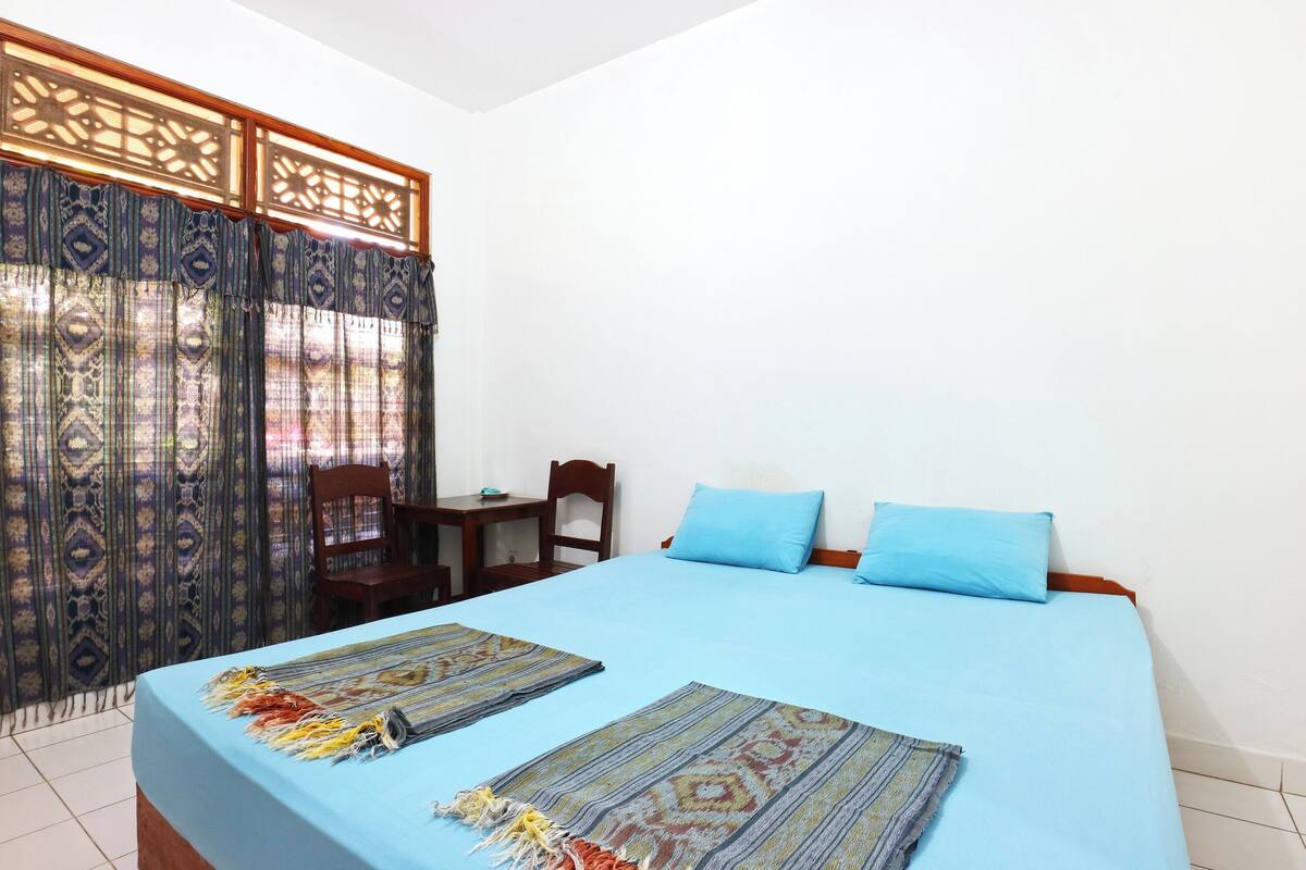 A bedroom with a bed, two blue pillows, a wooden headboard, a small table, and a chair.