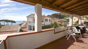 A covered balcony with white chairs and a table, overlooking a coastal town with terracotta roofs and a clear sky.
