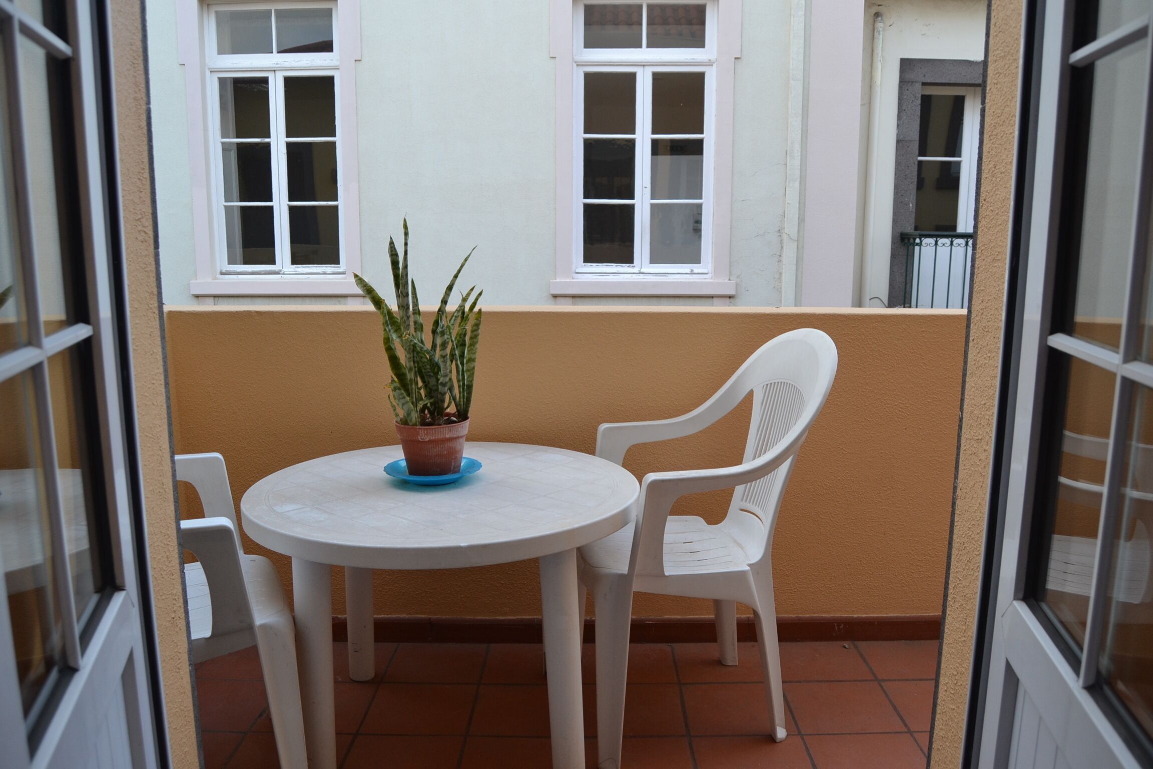A small balcony with a round table and two white chairs, a potted plant, and two windows.