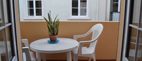 A small balcony with a round table and two white chairs, a potted plant, and two windows.