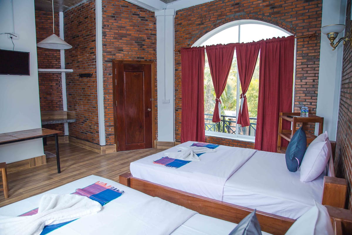 Ein Zimmer mit zwei Betten, einem Holztisch, einem Stuhl und einem Fenster mit roten Vorhängen.