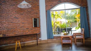 Ein Zimmer mit Backsteinwand, Holzboden, einem großen Fenster mit Vorhängen, einer hängenden Leuchte und einem kleinen Tisch mit Stühlen.