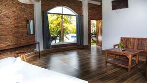 Ein Zimmer mit Backsteinwand, Holzdielenboden, einem großen Fenster mit Vorhängen, einer Couch, einem Tisch und einem an der Wand befestigten Fernseher.