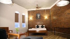 Ein Zimmer mit einem Bett, einem Holztisch und einem blauen Sofa. Es gibt zwei Fenster mit Rundbögen, eine Ziegelwand und einen Deckenventilator.