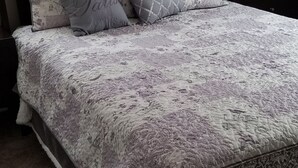 Siististi pedattu sänky kuviollisilla vuodevaatteilla, yöpöydällä oleva lamppu ja yöpöytä, jossa on kello.